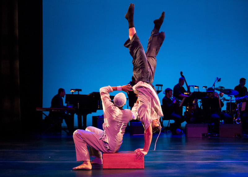 A man sits on a wooden box as another dancer does a hand stand off of it. The band is in the background.